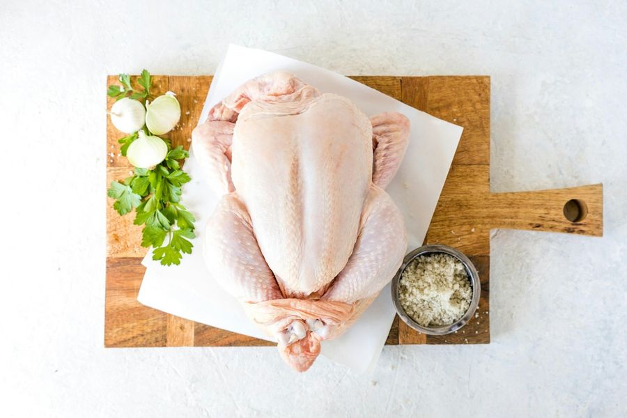 A whole raw chicken on parchment on a wooden board, with parsley, onions and salt